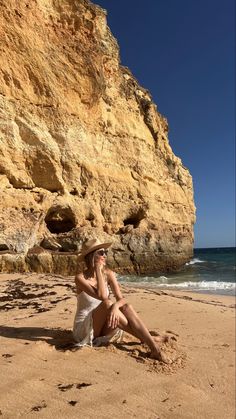 a woman sitting on top of a sandy beach next to the ocean wearing a hat