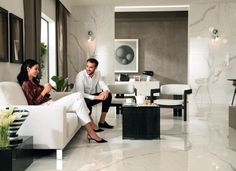 a man and woman sitting on couches in a living room with marble walls, flooring and large windows