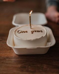 a birthday cake with a candle that says one year on it sitting in a white container