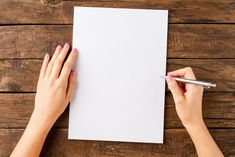 two hands holding a pen and writing on a piece of paper next to a wooden table