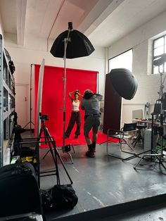 two people are standing in front of red backdrops and lighting equipment while one person is taking a photo