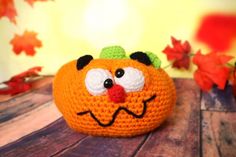 an orange crocheted pumpkin with eyes and nose sitting on a wooden table next to leaves