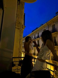 two women standing on a balcony at night drinking from wine glasses in front of them