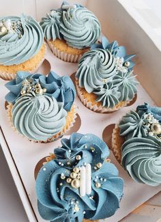 cupcakes with blue frosting and gold decorations in a white box on a table