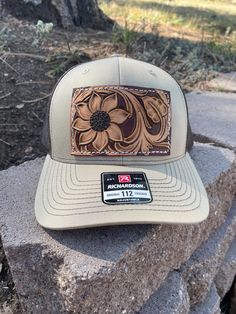 A tan and brown Richardson Trucker hat decked out with a sunflower and some vines on a leather patch! These Richardson  hats are super comfortable and have an adjustable snap back.  PLEASE READ: This item is for pre-ordering, and is made to order! Once purchased, this item will take approximately two weeks to re-create. Item will be a close replica, but each one will have slight variances in color, carving, etc. as I am not perfect, and leather is not perfect either! *All of HCL's leather products are cut, designed, dyed, painted, sewn, and assembled by hand. Items will not be machine perfect, but I try my hardest to get them there! Please keep in mind that leather may have natural scuffs, brand marks, discoloration, etc. It's all part of what makes leather so beautiful and unique! Brown Western Trucker Hat With Flat Brim, Western Style Brown Trucker Hat For Outdoor, Western Style Brown Trucker Hat With Flat Bill, Western Style Brown Snapback Hat, Western Brown Snapback Hat, Brown Trucker Hat For Western-themed Events, Brown Western Style Baseball Cap, Western Brown Hat With Leather Patch, Western Style Brown Hat With Leather Patch