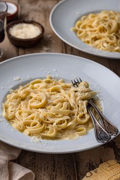 two white plates with pasta and cheese on them next to wine glasses, napkins and utensils