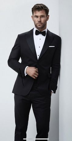 a man in a tuxedo leaning against a wall with his hands on his hips