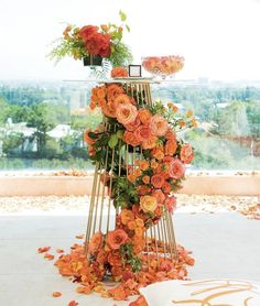 an arrangement of orange flowers and greenery on a metal stand in front of a window