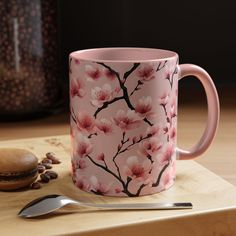 a pink coffee mug sitting on top of a wooden table next to a spoon and cup