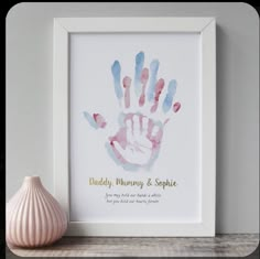 a pink and blue hand print on a white frame next to a vase with a pink flower