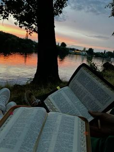 an open book sitting on top of a grass covered field next to a river at sunset