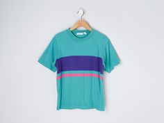 This vintage 90s t-shirt has one pink & one purple stripe. The tag size is L - it measures 21" across the chest laid flat, and it measures 24" long shoulder-to-hem. This item is in excellent vintage condition.   Content + Care - 100% cotton - Machine wash warm. Tumble dry low. Size + Fit - Tag size is L   - Chest: 21" across    - Length: 24" long shoulder-to-hem Green 90s Style T-shirt For Summer, Sporty Striped Summer T-shirt, 90s Striped Short Sleeve Tops, Retro Purple Crew Neck Top, 90s Striped Crew Neck Top, Green Short Sleeve T-shirt With Three Stripes, Striped 90s Style T-shirt For Summer, Striped 90s Style Summer T-shirt, 90s Striped T-shirt For Summer