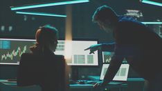 two people in front of three computer screens, one pointing at the screen while the other points