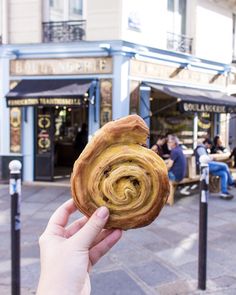 a person holding up a pastry in front of a building with people sitting at tables