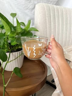 a person holding a glass cup with liquid in it on a table next to a potted plant