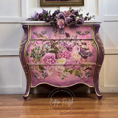 an old dresser painted in pink and purple with flowers on the top, next to a painting