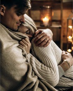 a man and woman wrapped up in blankets looking at each other with christmas lights behind them