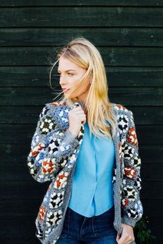 a woman standing in front of a black wall wearing a blue shirt and jeans with an open jacket over her shoulders