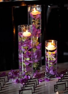 three tall clear vases filled with purple flowers and lit candles on a white table