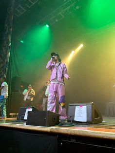 a man standing on top of a stage in front of green and yellow lights with his hands up to his head