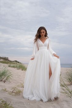 a woman standing on top of a sandy beach wearing a white dress and long sleeves