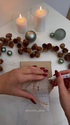 two hands with red nail polish are holding a piece of paper next to some pine cones and candles