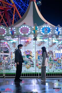 a man and woman standing in front of a merry go round ride at night time