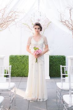 a woman in a wedding dress standing under a white tent holding a bouquet of flowers