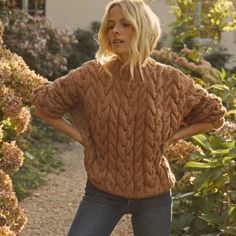 a woman is standing in front of some bushes and flowers wearing a brown cable knit sweater