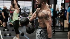 a man with no shirt holding a dumbbell in a gym while other people are doing exercises behind him