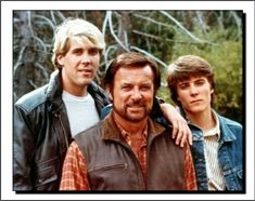 three young men standing next to each other in front of some trees and bushes with one man wearing a leather jacket