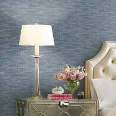 a nightstand with a lamp and flowers on it in front of a blue wallpaper