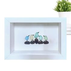 three little birds sitting on top of rocks in a white frame next to a potted plant