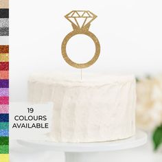 a white cake with a gold ring on top and rainbow glitters in the background