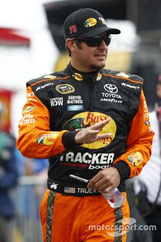 a man in an orange and black racing suit talking to someone on the sidelines