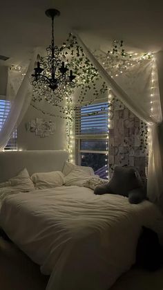 a white bed sitting under a chandelier in a bedroom next to two windows