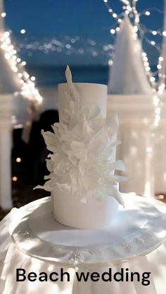 a white wedding cake sitting on top of a table next to the ocean with lights