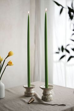 two green candles sitting on top of a table next to a vase with yellow flowers