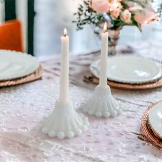 two white candles are sitting on a table with plates and flowers in the vase behind them