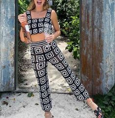 a woman standing in front of an open gate holding a drink and wearing black and white patterned pants