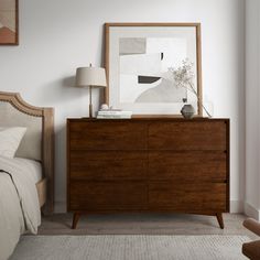 a bedroom scene with focus on the bed and dresser in the foreground as well as an art piece