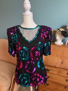 This listing is for a Leslie Fay Evenings short sleeve sequined black silk blouse. The sequins are pink, purple, and turquoise and are in a lovely floral design. There is a sheer mesh area on front of blouse. The blouse is 100% silk and lining is 100% polyester. Made in India. It is labeled a size small. It has shoulder pads and a back zipper. **Please note there is corrosion on hook/eye** It is missing eye at top of zipper.  Care Instructions: Dry Clean Condition: Good, used condition. There is some corrosion/rust on hooks/eyes but otherwise no damage. Size: Labeled Size Small. Please see lay flat measurements below. Lay Flat Measurements: Pit to Pit: 17.5 inches Wasit: 31 inches (doubled) Approximately 24 inches long from shoulder to hem Mannequin is size: 15" shoulder, 26 and 7/9" waist Glamorous Short Sleeve Blouse, Fitted Purple Sequin Top, Glamorous Short Sleeve Summer Blouse, Glamorous Embellished Short Sleeve Blouse, Glamorous Short Sleeve Embellished Blouse, Purple Short Sleeve Top For Party, Pink Short Sleeve Blouse For Party, Short Sleeve Blouse For Party Season, Embellished Short Sleeve Tops For Party Season