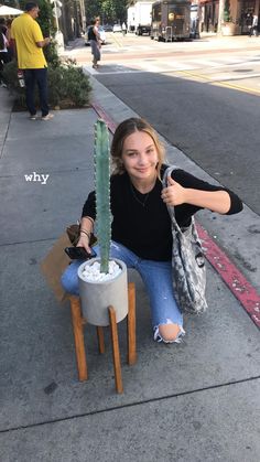 a woman sitting on the sidewalk next to a potted cactus with her thumb up