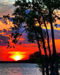 the sun is setting behind some trees on the water's edge with clouds in the sky