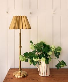 a vase with green flowers and a gold lamp on a wooden table in front of a white wall