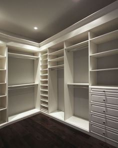 an empty walk in closet with white shelving and drawers on the wall, along with dark wood flooring