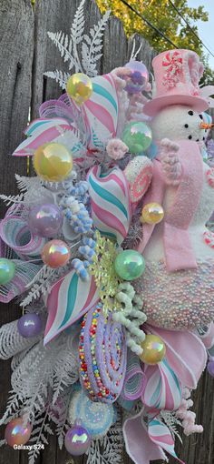 a pink and white christmas wreath with a snowman on it's head next to a wooden fence