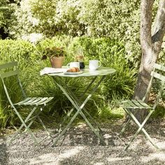 two green chairs and a table with food on it in front of some trees, grass and bushes