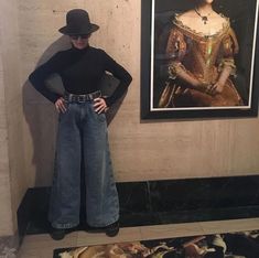 a woman standing in front of a painting wearing jeans and a black hat with her hands on her hips