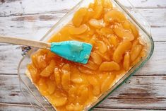 a glass dish filled with sliced peaches on top of a white wooden table next to a blue spatula